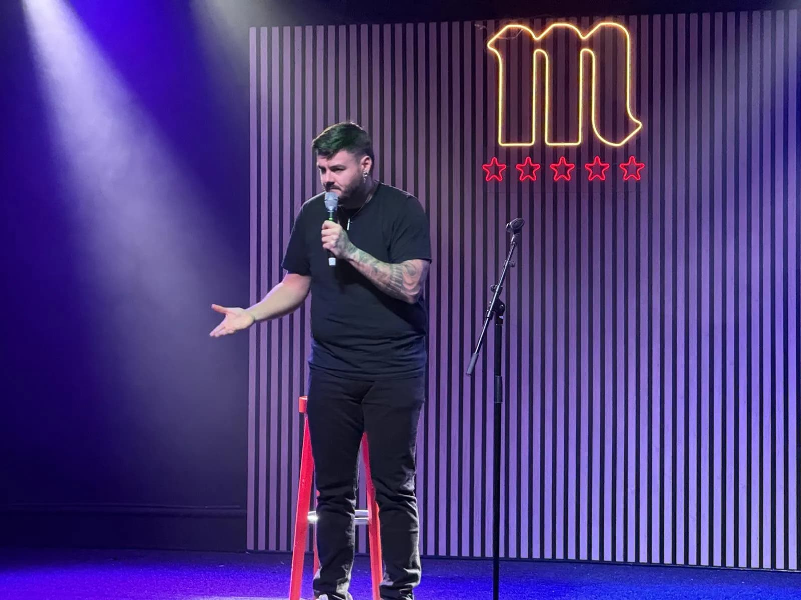 Juanjo Albiñana with the microphone on the La Bohemia stage, lit in purple in front of an illuminated sign with an «m» and five stars