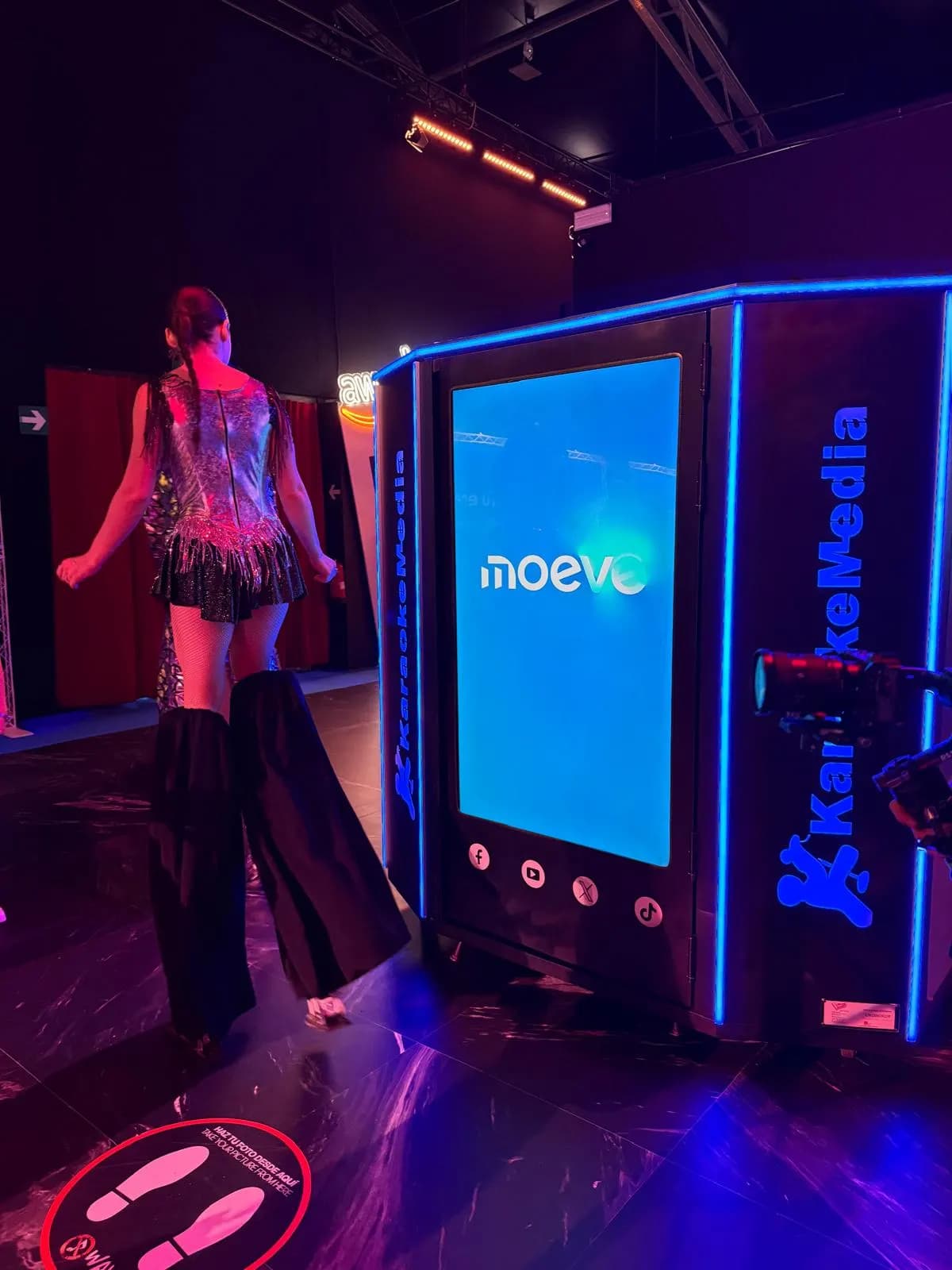 Artista amb xanques i vestit brillant al costat de la cabina de karaoke amb el logo de Moeve a la pantalla