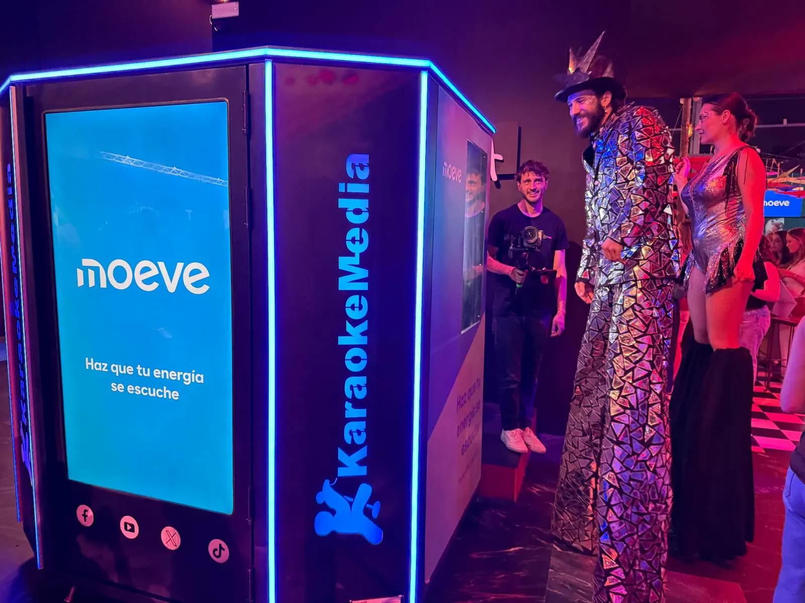 Cabina de karaoke amb llum blava i la pantalla exterior amb el logo de Moeve i el lema «Haz que tu energía se escuche», al costat de dos artistes amb xanques a IFEMA