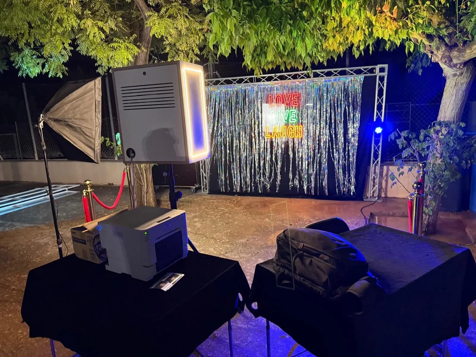 Fotomatón con marco de luz y foco de estudio delante de un photocall con cortina plateada y el neón «LOVE LIVE LAUGH», con la impresora en primer plano