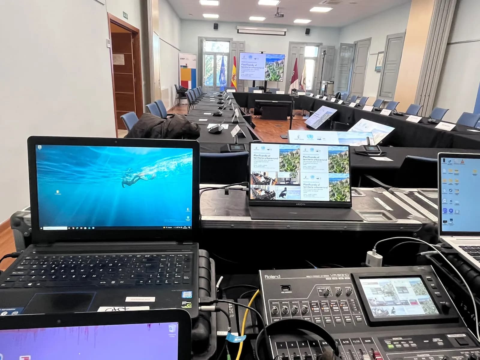 Control desk with laptops, a monitor showing several signals at once and the video switcher, facing the prepared room