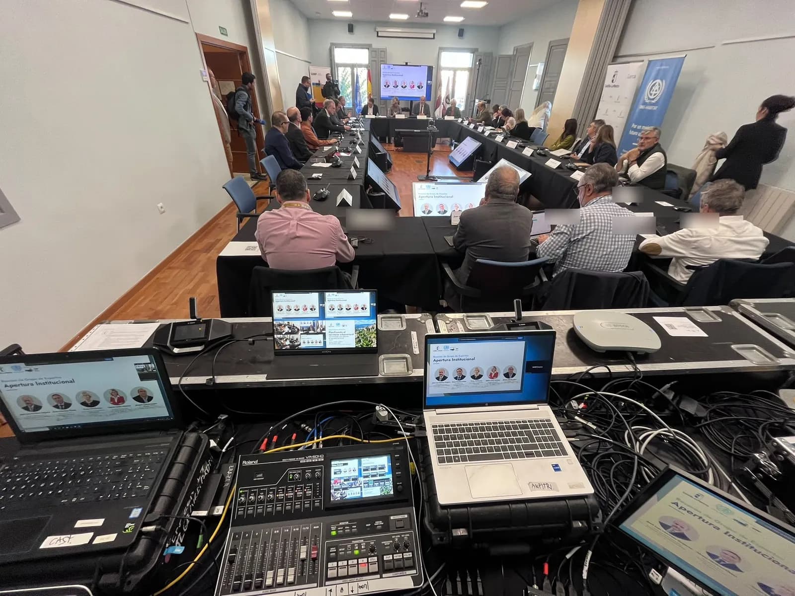 The meeting under way seen from the control desk, with the video switcher and laptops in the foreground and the participants, seen from behind, around the U-shaped table